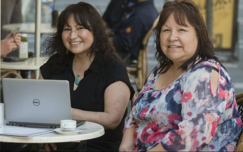 Three indigenious women smiling and sitting in front of a laptop in a cafe setting.